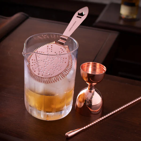 Cocktail Kingdom Seamless Paddle Mixing Glass Tapered Base shown with a gold jigger and gold mixing spoon on a bar setting.