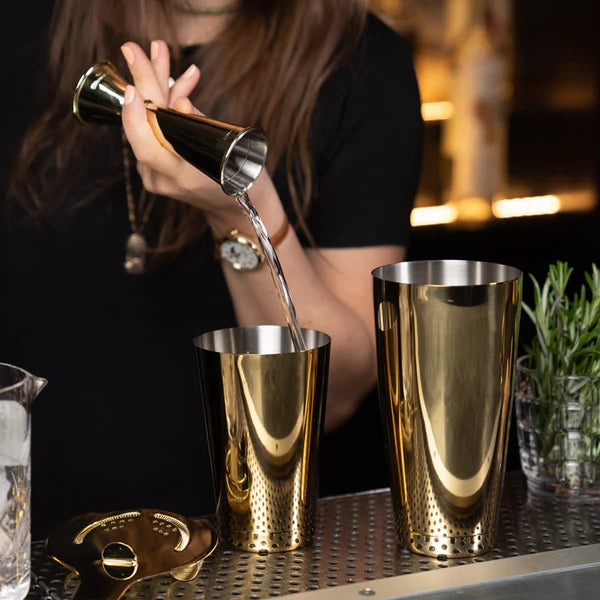 1121 G Gold Cocktail Kingdom Koriko Set of 2 Weighted Tins, showing cocktail being poured in by a gold jigger with a gold strainer and mixing glass on bar top.
