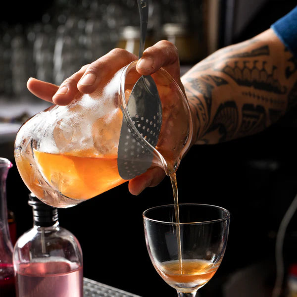 Cocktail Kingdom Premium Julep Strainer in stainless steel shown straining cocktail into glass in dark moody bar background.