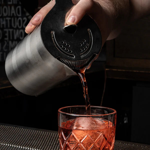 1126 Cocktail Kingdom Koriko Hawthorne Strainer shown pouring cocktail from shaker tin into a mixing glass on a dark moody bar background. 