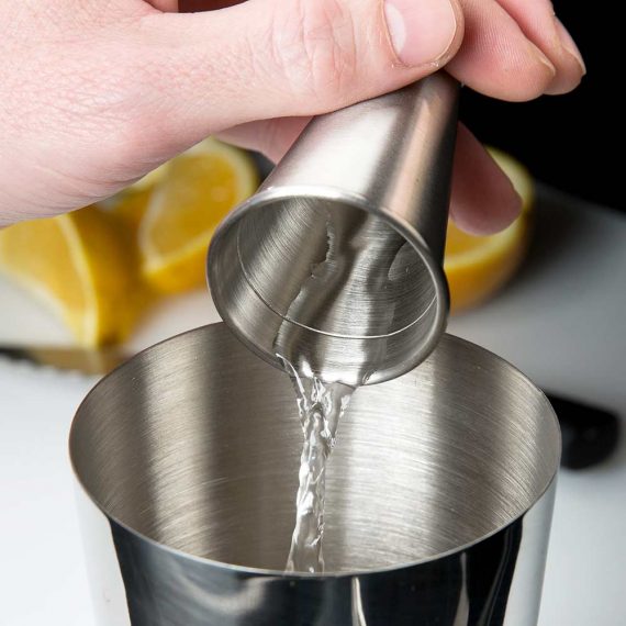 Metallic jigger pouring liquid into a cocktail shaker with lemons in the background