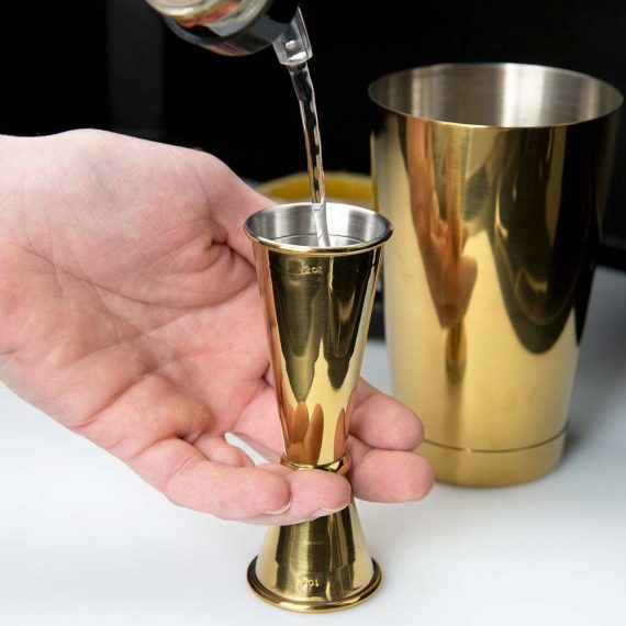 Gold jigger being filled with liquid with gold shaker in the background shot on white foreground.