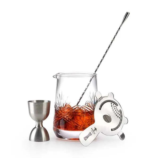 Barfly Wide Base Mixing Glass 25 ounces / 740 ml shown filled with liquid, being stirred by barspoon with silver jigger and silver cocktail strainer beside it on white silo background.