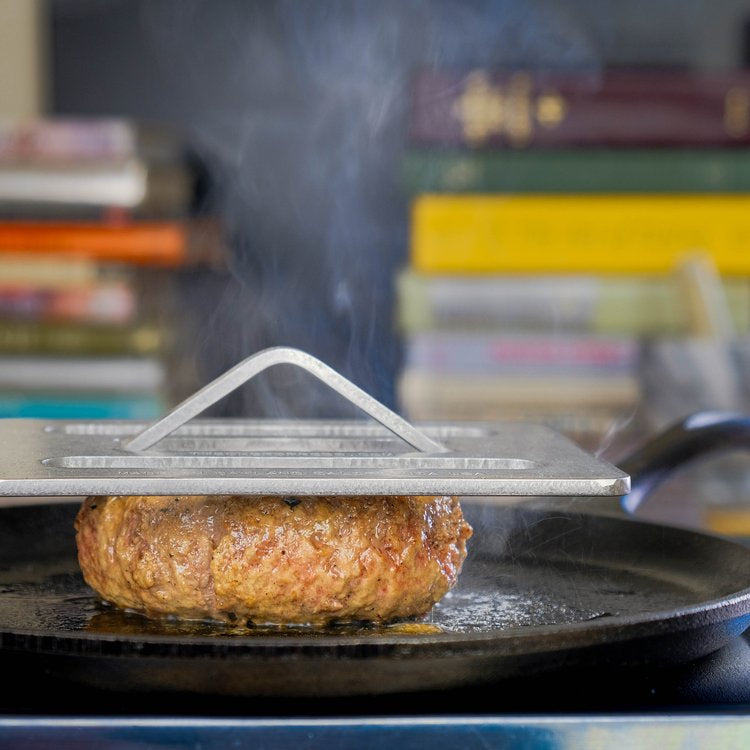 Hamburger patty cooking on a grill with a Chef press above it, books in the background.