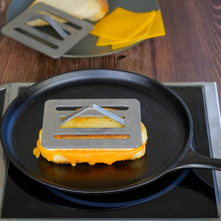 Grilled cheese sandwich being pressed in a pan with a Chef press on a wooden surface.