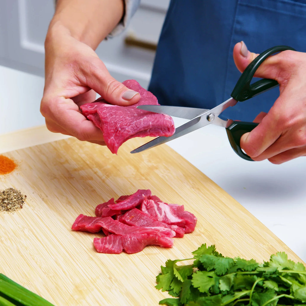 Joyce Chen Scissors in dark navy shown cutting meat on cutting board.