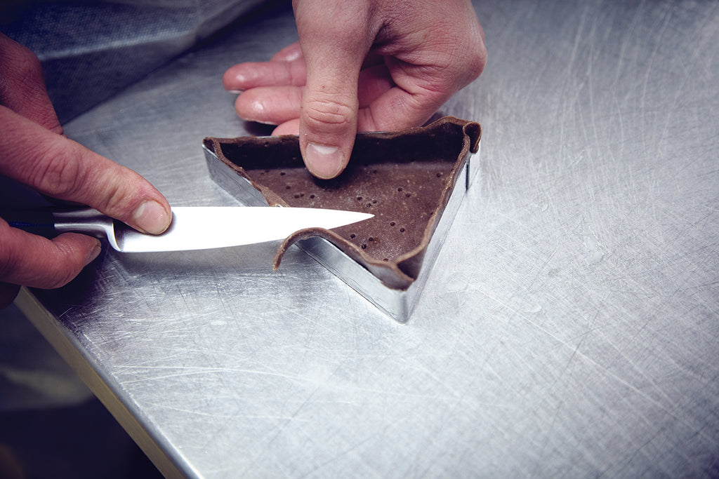 Person cutting a triangular piece of chocolate with a knife, from a triangular tart mold on a metal surface.