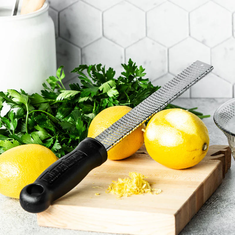 u746 black microplane Zester on a cutting board with lemons and herbs on a kitchen counter.