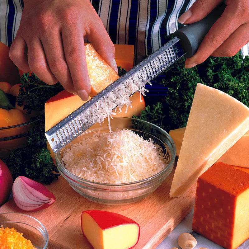Person grating cheese into a bowl with U746 Microplane classic zester with various cheeses and vegetables surrounding on a wooden board.