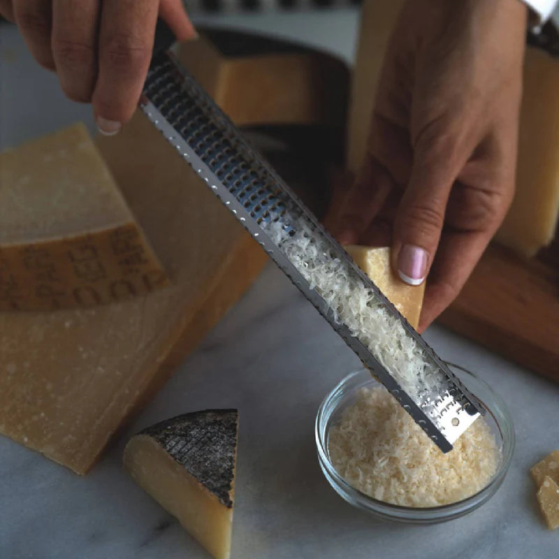 Person using a class microplane grater to grate Parmesan cheese into a small glass bowl with more cheese and a block of cheese in the background.