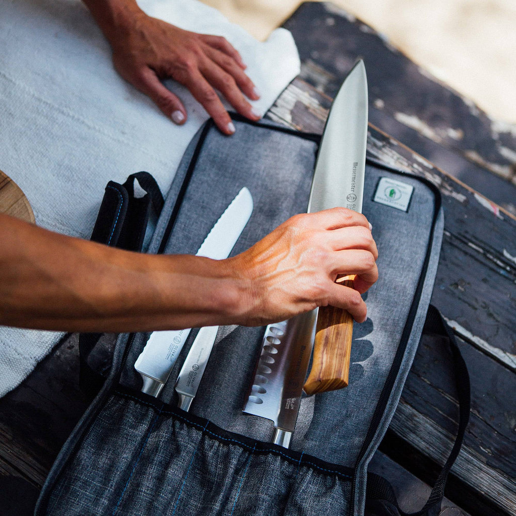 Person holding a knife over a set of knives on a gray knife roll with a wooden surface in the background.