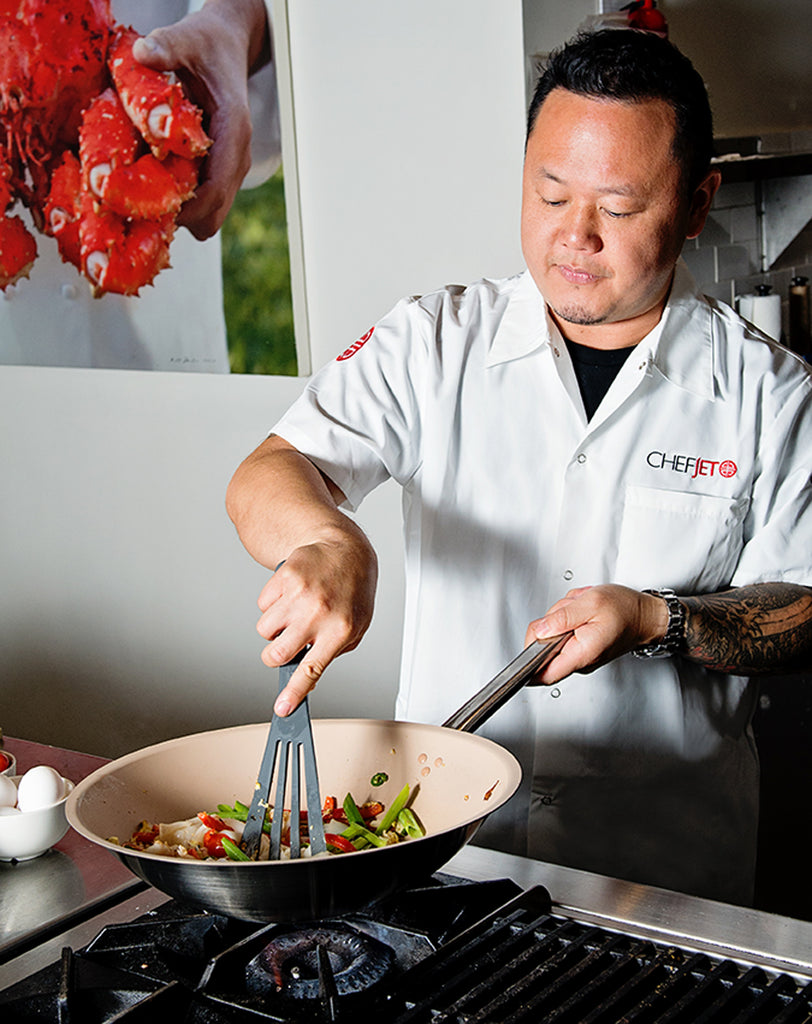 Chef cooking in a kitchen with a wok and vegetables, wearing a white chef's coat using grey slotted spatula.