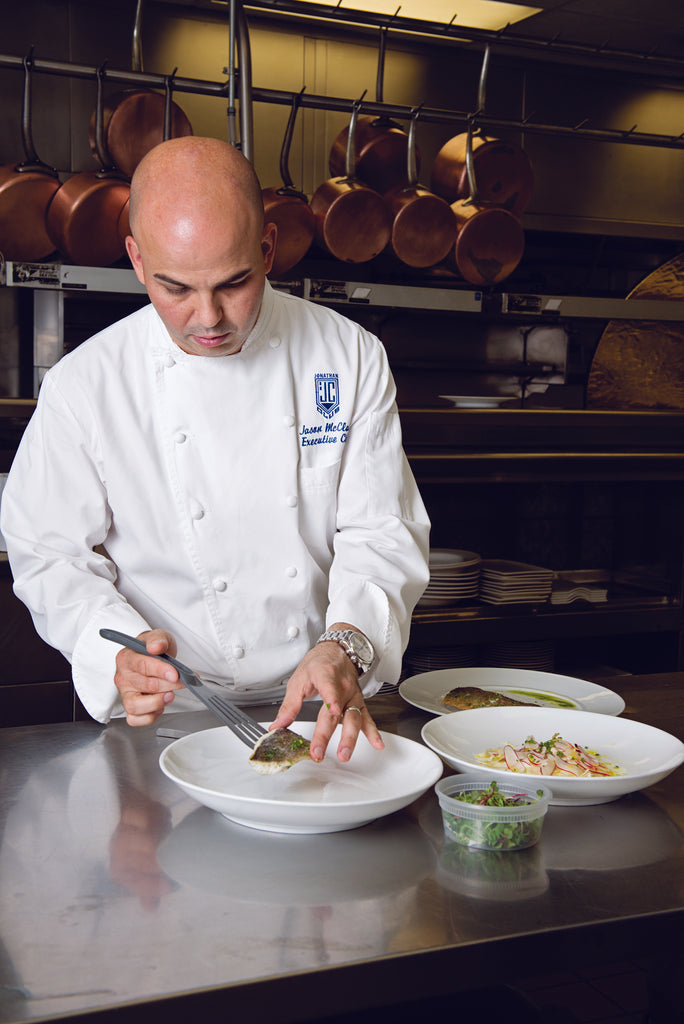 Chef in a kitchen plating food with grey slotted spatula with pots hanging in the background
